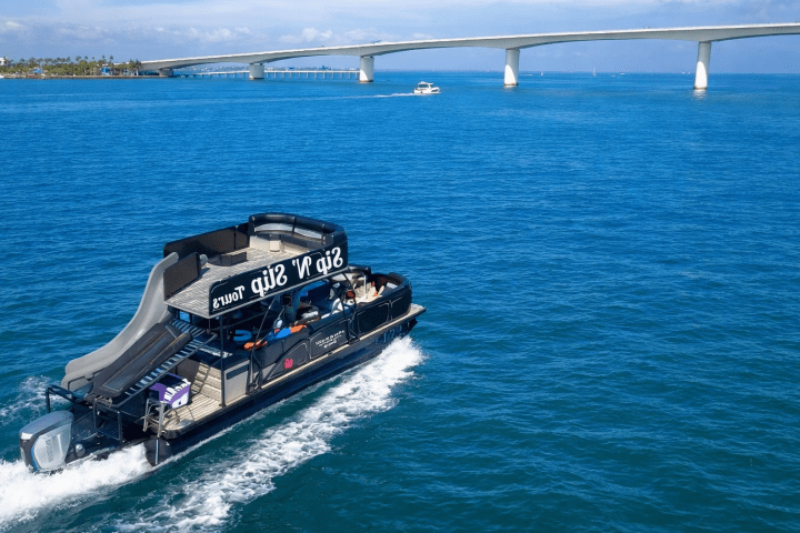 Tour boat with slide on blue water near a long bridge under a clear sky.