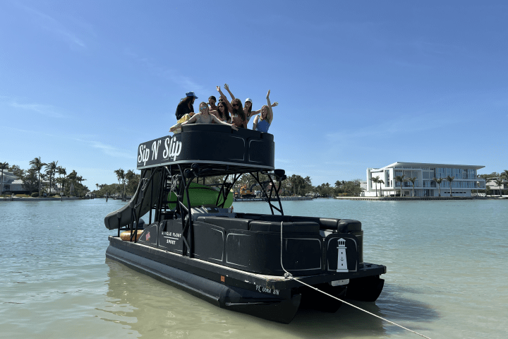 Group of people on a double-deck pontoon boat named 'Sip N' Slip' in a sunny waterway.