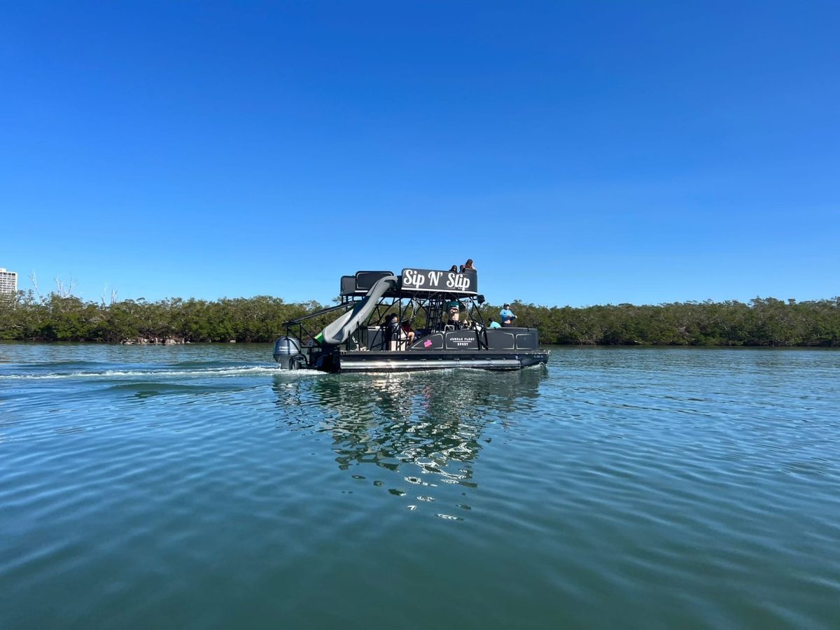 Party boat with slide on calm water, clear sky, trees in the distance.