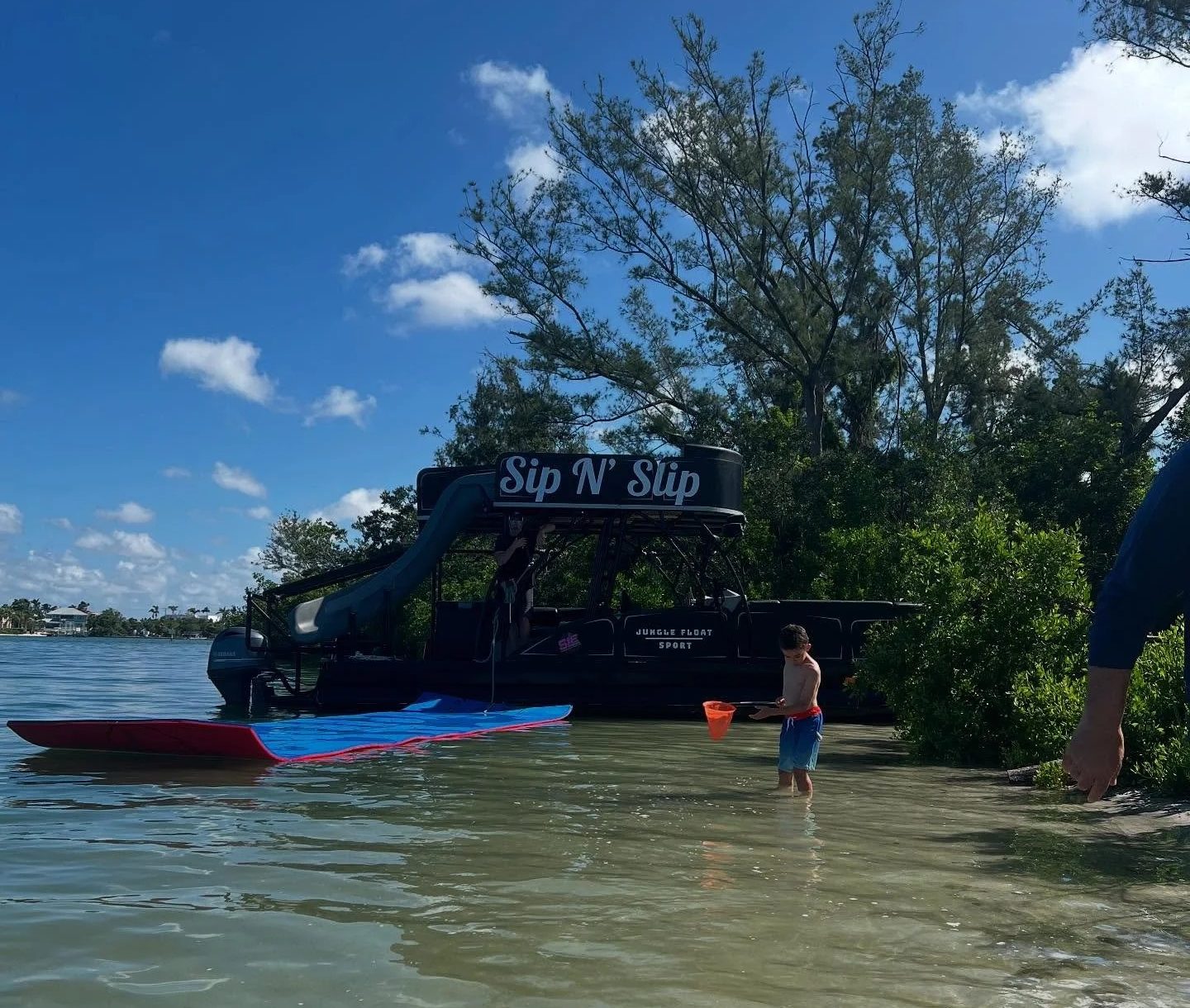 Child with red bucket by water, slide with 'Sip N' Slip' sign, trees and blue sky in background.